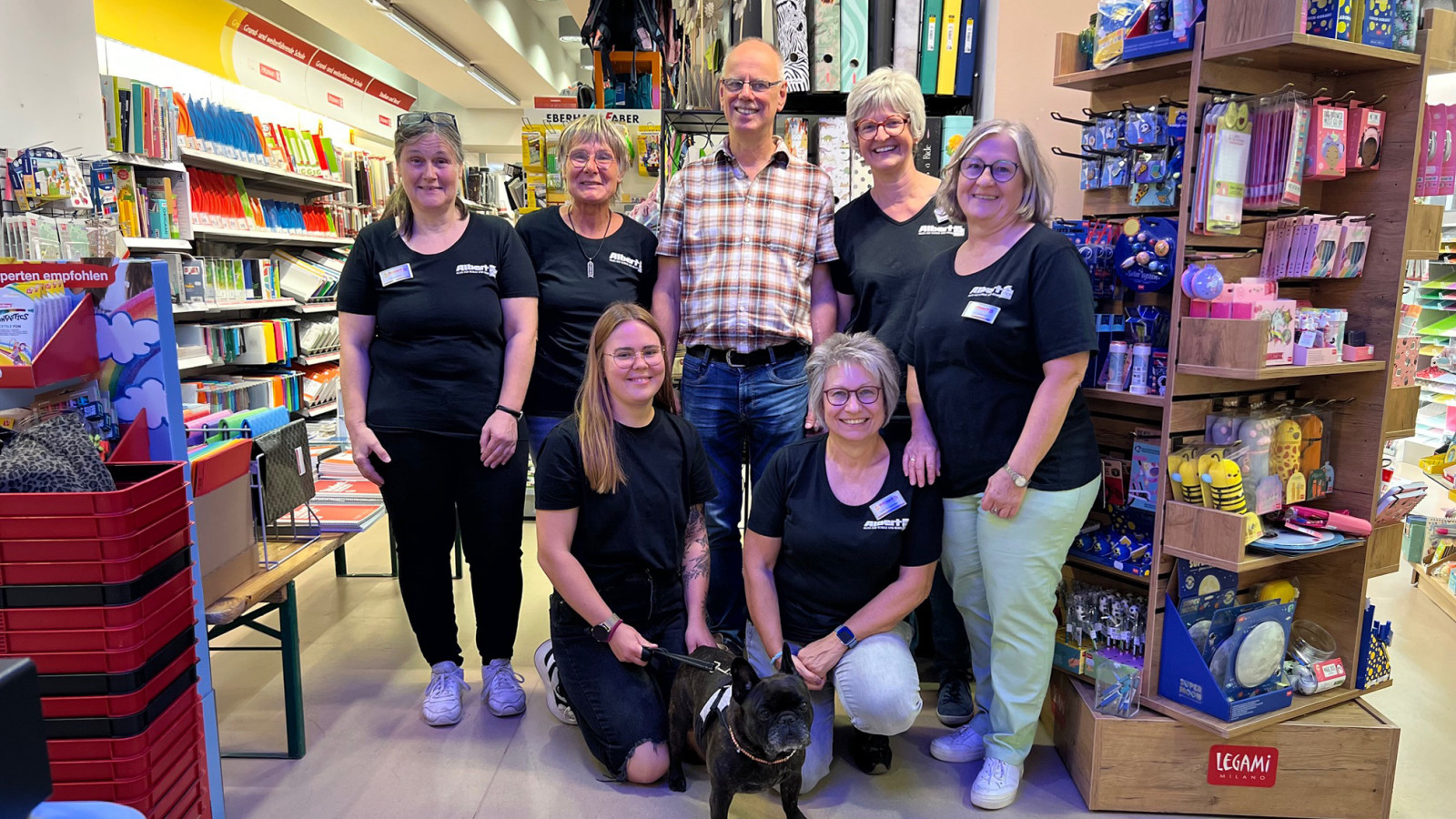 Team aus Menschen und einem Hund in einem Schreibwarengeschäft vor Regalen mit Büroartikeln.