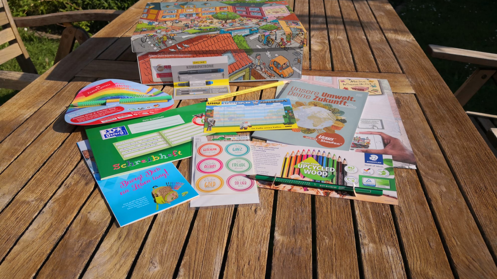 Lernmaterialien wie Bücher, Stifte, Karten und ein Spielkasten auf einem Holztisch im Garten.
