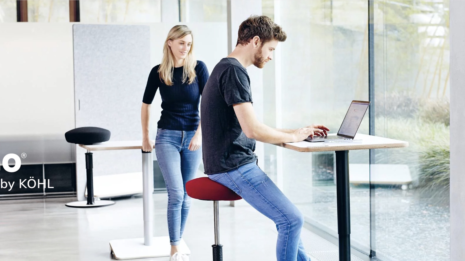 Mann und Frau in modernem Büro an höhenverstellbaren Tischen mit dynamischen Sitzgelegenheiten.