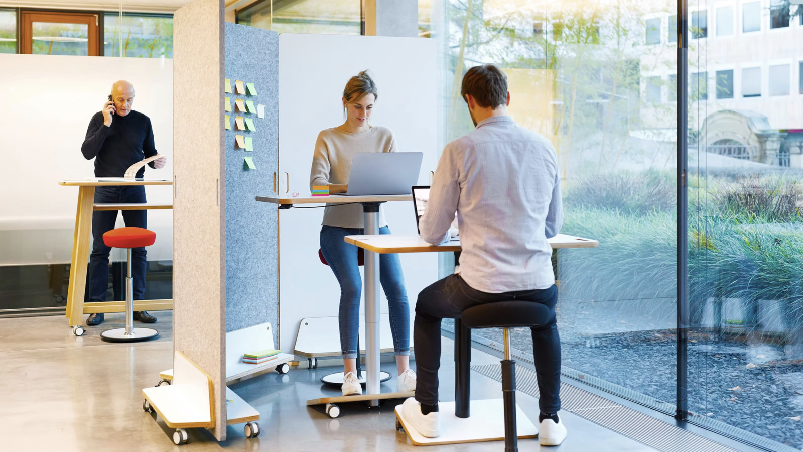 Drei Personen arbeiten in einem modernen Büro an Stehpulten, umgeben von großen Fenstern.