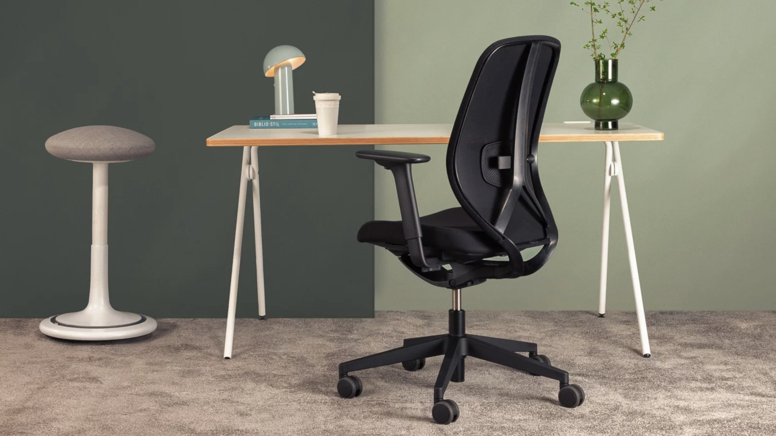 Büroeinrichtung mit schwarzem Stuhl, Schreibtisch, Lampe und Vase auf grauem Teppich.