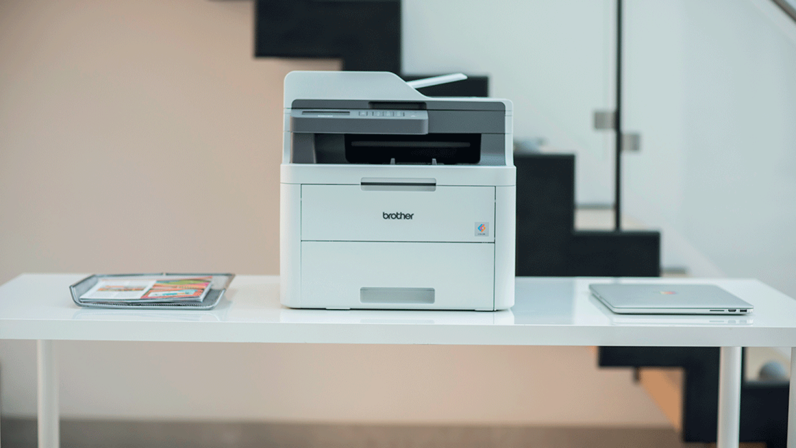 Drucker auf einem weißen Tisch mit Laptop und Tablett, Hintergrund zeigt Treppen.