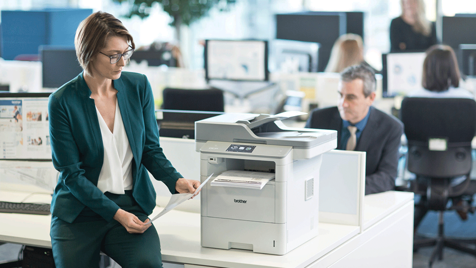 Büroangestellte prüft Dokumente neben einem Drucker, Kollegen arbeiten im Hintergrund an Schreibtischen.