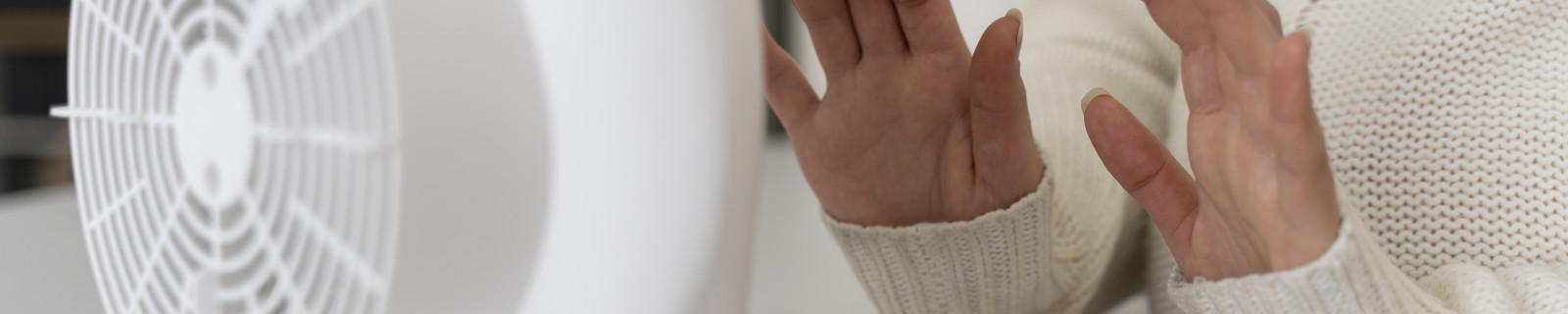 Frau in beigem Pullover wärmt Hände an einem weißen Heizgerät auf einem Tisch.