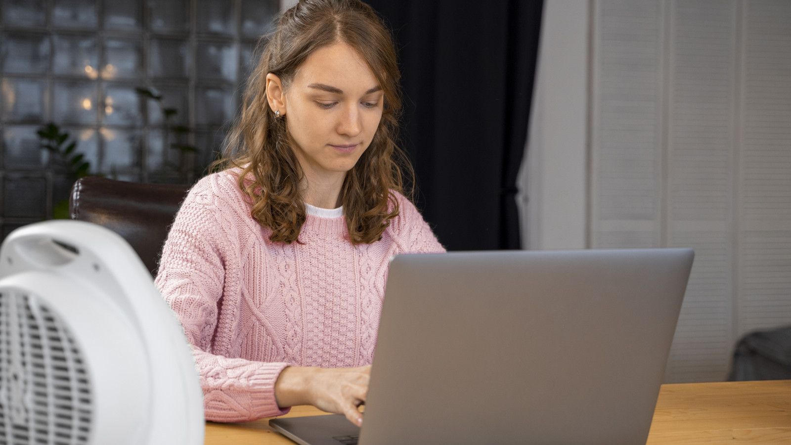 Frau im rosa Pullover tippt auf einem Laptop, mit einem Heizlüfter im Vordergrund.