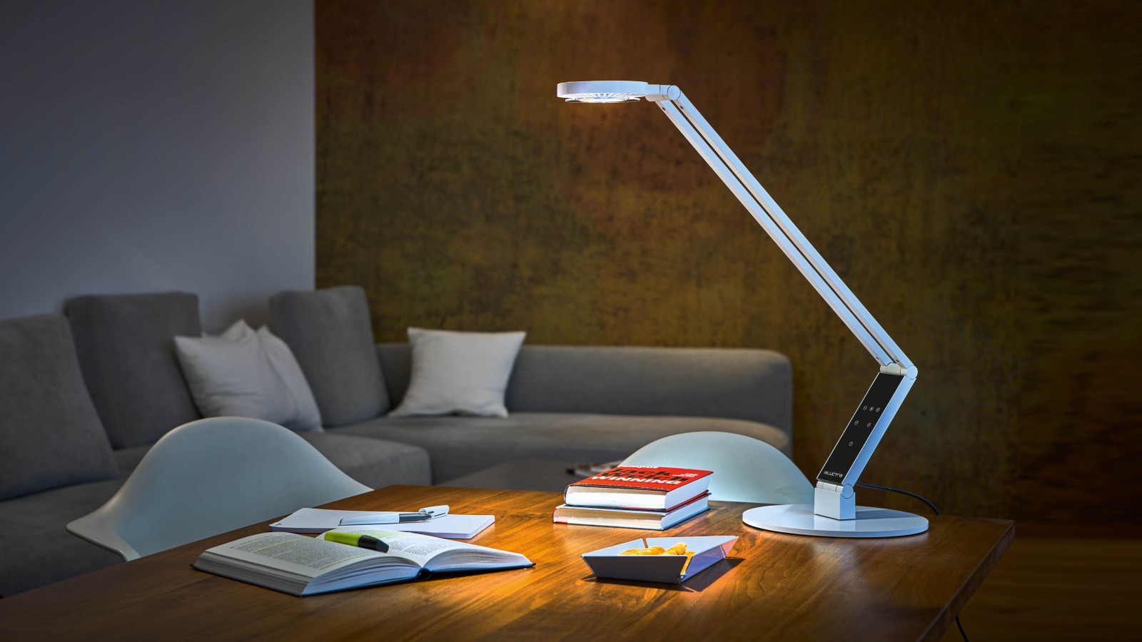 Schreibtisch mit LED-Lampe, Büchern, Notizbuch und Snackschale im Wohnzimmer.