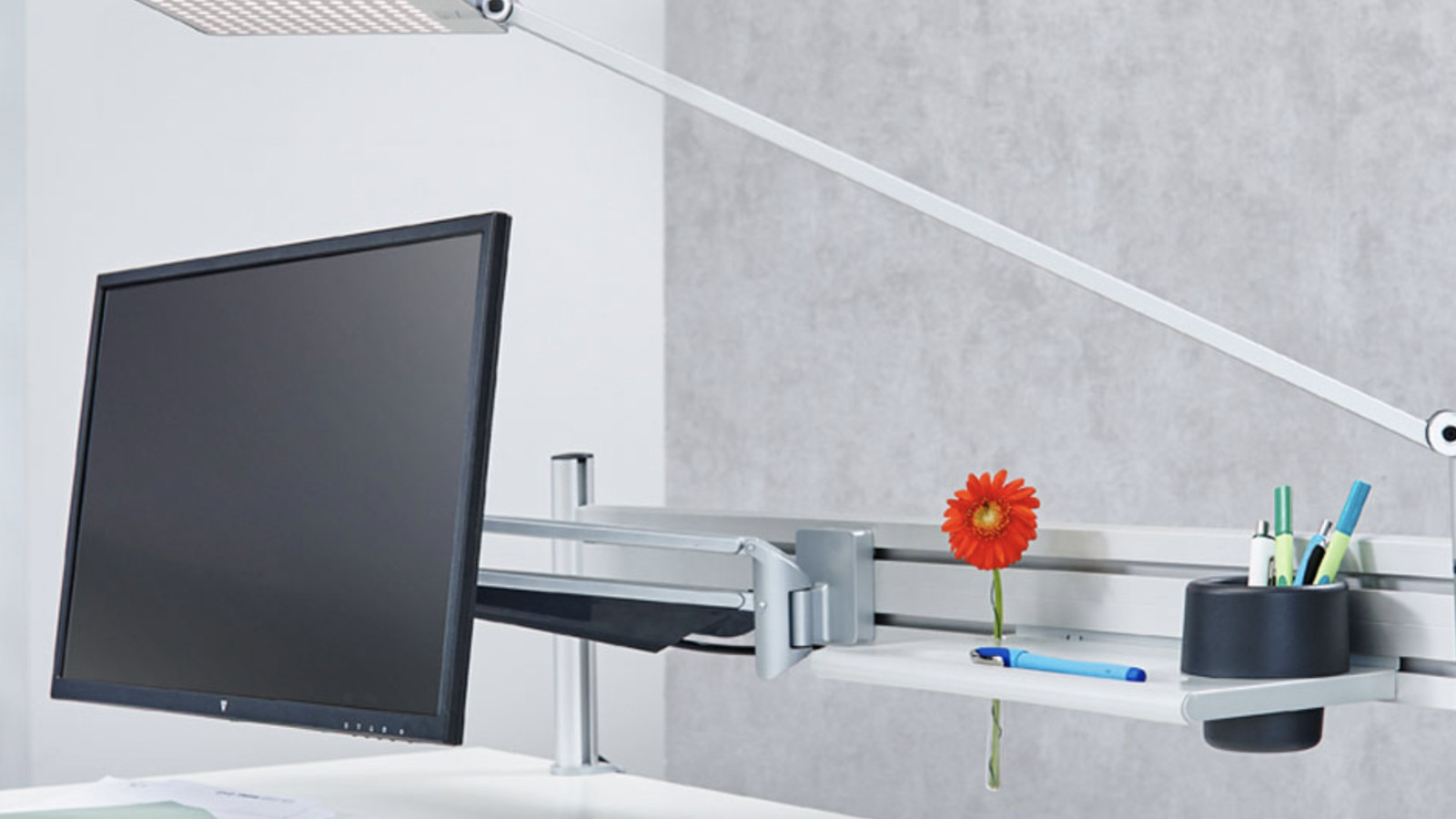 Arbeitsplatz mit Bildschirm, Lampe und einer roten Blume in der Halterung.