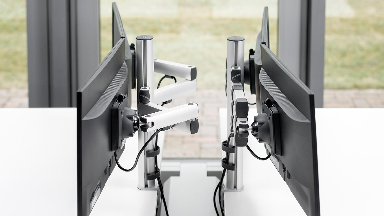 Zwei Monitore auf Schreibtischen mit Monitorarmen vor einem Fenster, Rückansicht.
